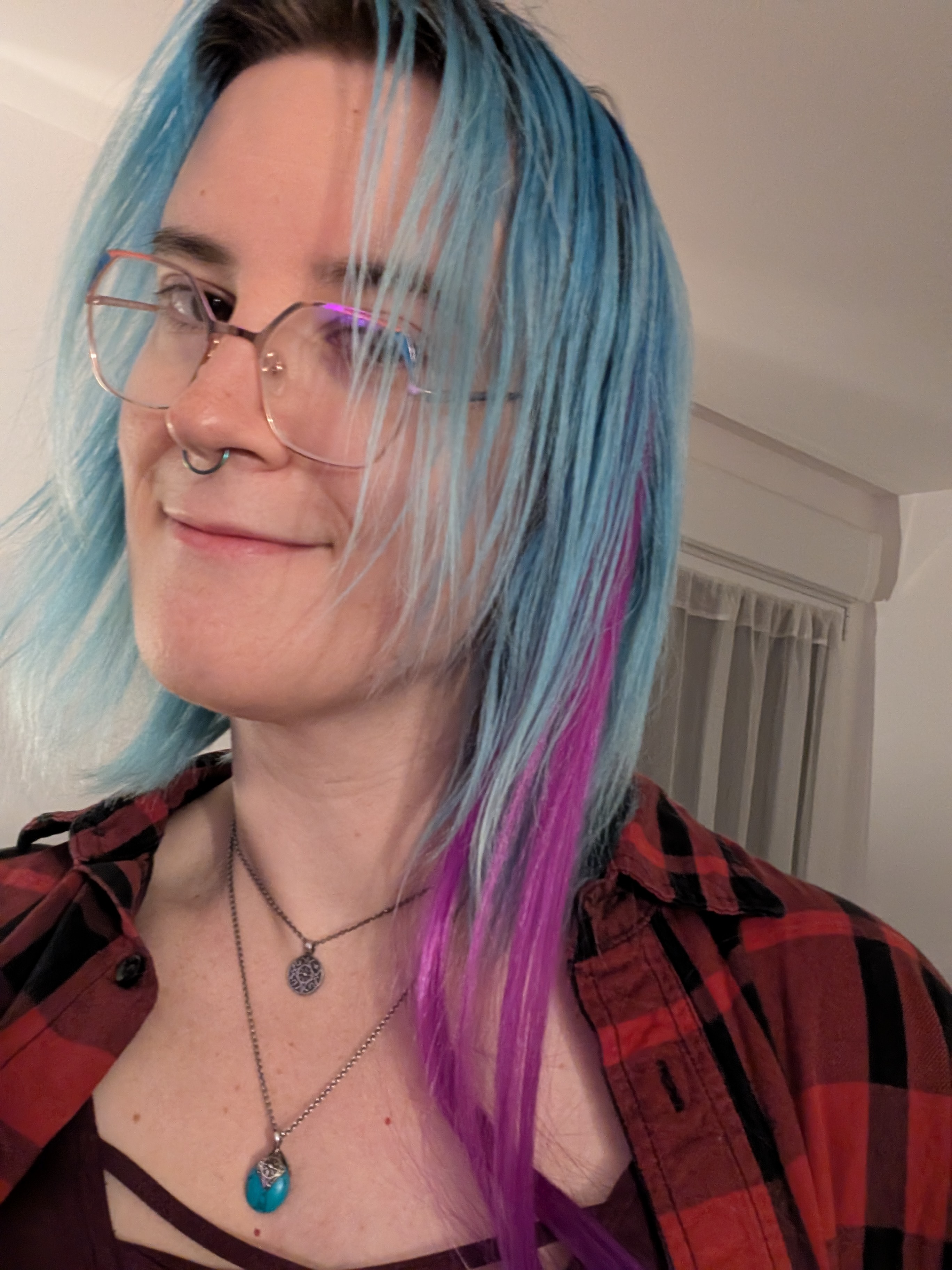 A white fem-looking person with blue hair (with a purple lock), slightly hexagonal blue and orange glasses and a septum ring is smiling, wearing a black dungaree over a dark pink top.